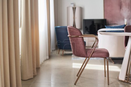 Photo for Soft velvet armchair of ash rose color standing by table in front of camera in modern furniture shop interior with beige curtains covering windows - Royalty Free Image