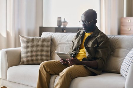 Foto de Joven afroamericano freelancer masculino con tableta sentado en sofá cómodo y suave en la sala de estar y mirando a través de datos en línea - Imagen libre de derechos