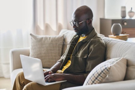 Photo for Young African American male entrepreneur in casualwear using laptop while sitting on soft comfortable couch in living room or office - Royalty Free Image