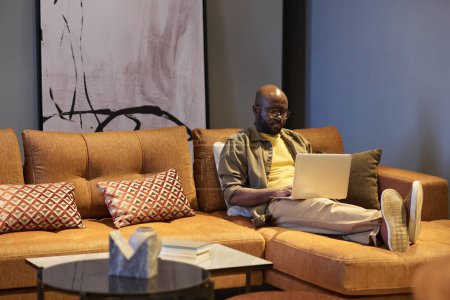 Téléchargez les photos : Sérieux jeune homme noir avec ordinateur portable sur les genoux assis sur un canapé confortable avec des coussins dans le salon tout en travaillant sur un nouveau projet - en image libre de droit