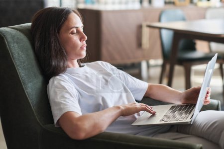 Téléchargez les photos : Jeune femme d'affaires sérieuse avec ordinateur portable assis dans un fauteuil confortable et regardant l'écran tout en regardant des vidéos en ligne ou en lisant des données - en image libre de droit