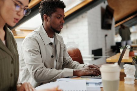 Téléchargez les photos : Focus sur le jeune analyste afro-américain tapant sur le clavier de l'ordinateur portable et regardant à travers les données en ligne à l'écran tout en étant assis par le lieu de travail - en image libre de droit