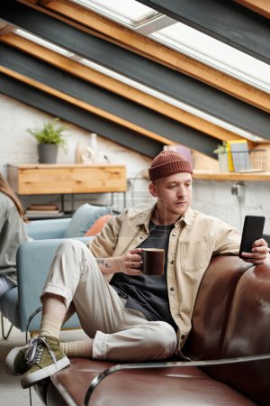 Foto de Joven hombre de negocios descansado en ropa de casualwear mensajes de texto en el teléfono móvil y tomar una taza de café mientras está sentado en el sofá de cuero delante de la cámara - Imagen libre de derechos