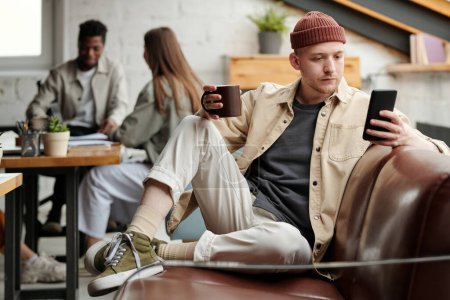 Foto de Joven trabajador de cuello blanco masculino serio en ropa de casualwear mensajes de texto en el teléfono inteligente o mirando a través de mensajes mientras está sentado en el sofá - Imagen libre de derechos