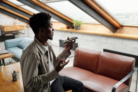 Photo for Side view of young African American male employee recording voice message in office while holding smartphone in front of his face - Royalty Free Image