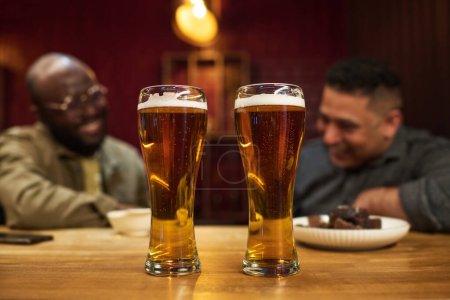 Téléchargez les photos : Deux grands verres de bière fraîche mousseuse fraîche avec de minuscules bulles à l'intérieur debout sur le comptoir de bar en bois contre deux copains interculturels - en image libre de droit