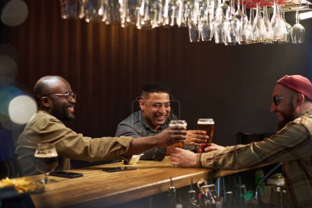 Téléchargez les photos : Barman barbu en lunettes de soleil, bonnet et chemise passant deux verres de bière à des copains masculins interculturels joyeux debout au comptoir du bar - en image libre de droit