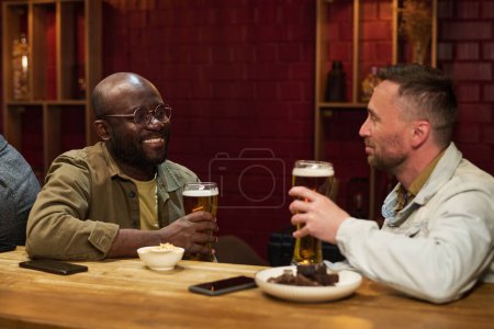 Photo for Happy young African American man with glass of beer looking at his buddy during conversation by bar counter while enjoying weekend - Royalty Free Image