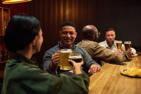 Téléchargez les photos : Un homme souriant avec une tasse de bière moussante grillant avec une jeune femme brune tout en restant debout près du comptoir du bar en bois et en profitant de la fête - en image libre de droit