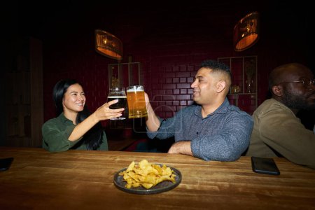 Photo for Happy young woman and her boyfriend clinking with glasses of beer while standing by bar counter with plate of potato chips and having nice time - Royalty Free Image