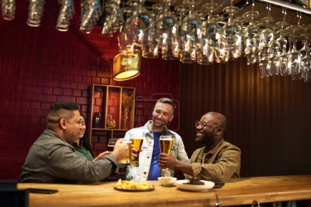 Foto de Grupo de alegres amigos interculturales brindando con vasos de cerveza mientras están de pie junto al mostrador del bar y disfrutando de la fiesta el fin de semana o después del trabajo - Imagen libre de derechos