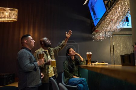 Foto de Vista lateral de jóvenes amigos interculturales tomando cerveza y viendo la emisión de hockey mientras uno de ellos expresa tensión y señala a la pantalla - Imagen libre de derechos