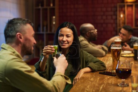 Photo for Happy young brunette woman with glass of aperitif toasting with her boyfriend while both sitting by bar counter and celebrating life event - Royalty Free Image