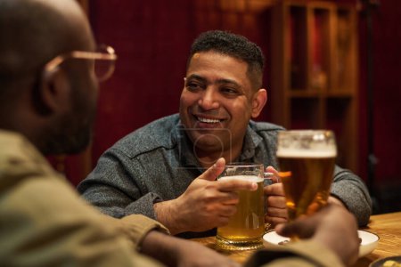 Foto de Concéntrate en un joven feliz con una taza grande de cerveza lager mirando a su amigo afroamericano durante el chat mientras ambos están sentados junto al mostrador del bar. - Imagen libre de derechos