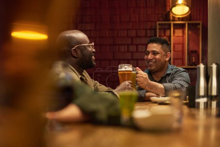 Foto de Dos jóvenes amigos interculturales felices tintinean con vasos de cerveza espumosa y se miran mientras están sentados junto al mostrador del bar - Imagen libre de derechos
