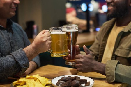 Foto de Manos de jóvenes hombres interculturales tintineando con vidrio y jarra de cerveza fresca y espumosa sobre la mesa mientras uno de ellos muestra el pulgar hacia arriba - Imagen libre de derechos