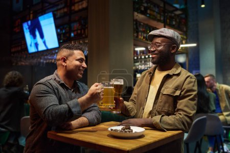 Foto de Dos amigos interculturales felices tintinean con un vaso y una taza de cerveza espumosa mientras se sientan a la mesa en el bar y disfrutan del fin de semana. - Imagen libre de derechos