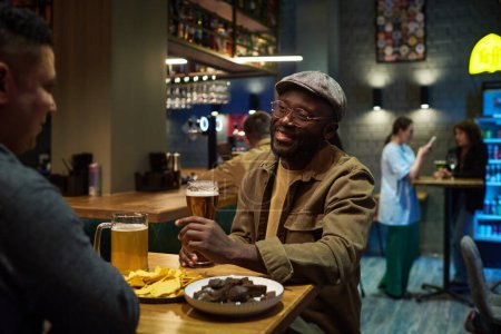 Foto de Feliz joven afroamericano hombre con un vaso de cerveza fresca hablando con su amigo mientras está sentado junto a la mesa frente a él contra el interior de la barra - Imagen libre de derechos