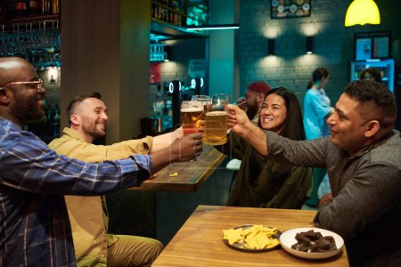 Téléchargez les photos : Groupe de quatre copains interculturels heureux clinking avec des tasses de bière sur la table en bois et comptoir de bar tout en étant assis dans un pub et avoir du bon temps - en image libre de droit