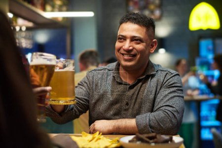 Foto de Sonriente hombre en ropa de casualwear mirando a su amigo mientras está sentado junto a la mesa en el bar el fin de semana y tintineo con taza de cerveza durante la tostada - Imagen libre de derechos