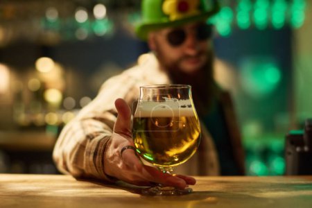 Foto de Concéntrese en la mano del barman barbudo con sombrero verde y camisa a cuadros poniendo un vaso de cerveza espumosa fresca en el mostrador de madera en el pub - Imagen libre de derechos