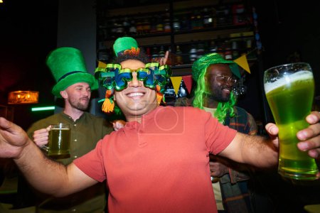 Foto de Joven hombre alegre en gafas de trébol animándose con un vaso de cerveza y bailando contra un grupo de amigos interculturales en la fiesta en el pub - Imagen libre de derechos