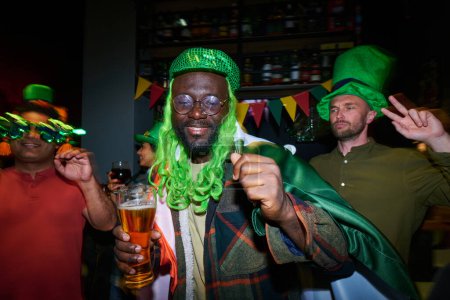 Foto de Joven feliz con peluca verde animándose con un vaso de cerveza y bailando contra amigos interculturales en la fiesta de San Patricio en el pub irlandés - Imagen libre de derechos