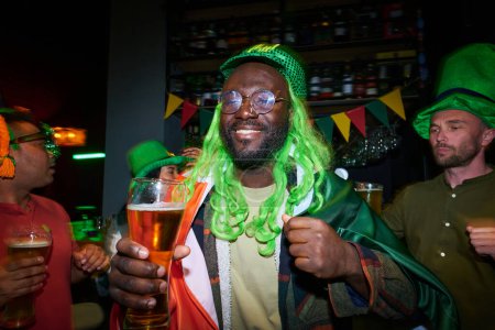 Foto de Feliz joven afroamericano con gorra verde y peluca bailando en la fiesta en el pub irlandés contra un grupo de amigos interculturales con vasos de cerveza - Imagen libre de derechos