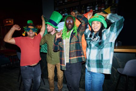 Foto de Grupo de jóvenes amigos emocionados en sombreros verdes bailando en la fiesta de San Patricio contra el mostrador del bar y mirando a la cámara mientras disfruta de la celebración - Imagen libre de derechos