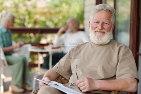Téléchargez les photos : Heureux homme âgé avec les cheveux blancs et la barbe assis dans le fauteuil devant la caméra et vous regardant tout en ayant du repos à la maison de retraite - en image libre de droit