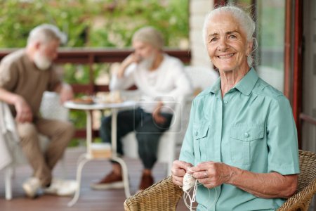 Photo for Cheerful senior woman with white hair looking at camera while sitting in wicker armchair on terrace of retirement house and knitting scarf - Royalty Free Image