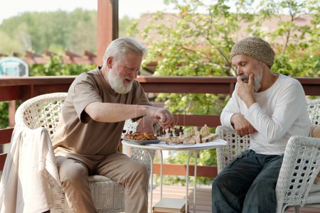 Téléchargez les photos : Deux hommes âgés barbus en tenue décontractée jouant aux échecs par une petite table ronde après le petit déjeuner assis dans des fauteuils sur le patio de la maison de retraite - en image libre de droit
