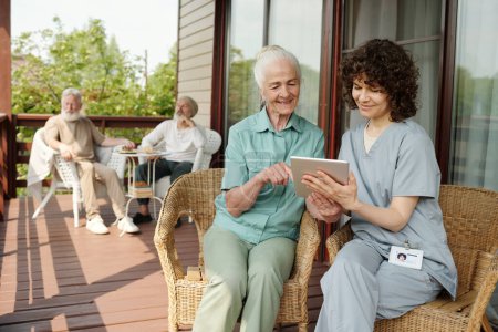 Téléchargez les photos : Aide soignant en uniforme montrant quelque chose à la femme âgée sur l'écran de la tablette et en discutant alors que les deux assis dans des fauteuils en osier sur le patio - en image libre de droit