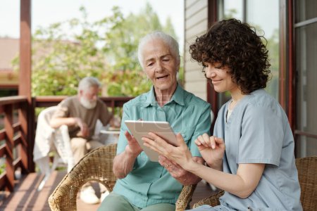 Foto de Enfermera joven feliz en uniforme haciendo presentación a paciente mayor de la casa de retiro mientras ambos miran la pantalla de la tableta - Imagen libre de derechos