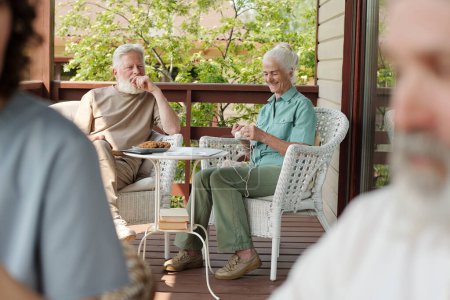 Photo for Focus on elderly couple sitting in wicker armchairs by small white table on terrace of retirement house while aged woman knitting something - Royalty Free Image