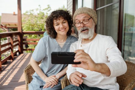 Photo for Happy young caregiver and senior man watching online movie or video in smartphone while sitting in armchairs on patio of retirement house - Royalty Free Image