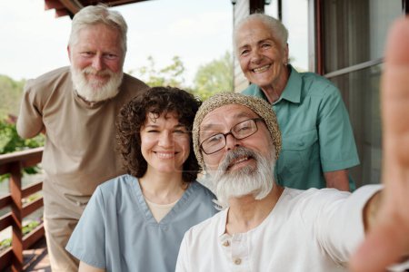 Foto de Feliz joven cuidador y grupo de personas mayores haciendo selfie en el teléfono inteligente en poder de uno de los pacientes varones ancianos de la casa de retiro - Imagen libre de derechos