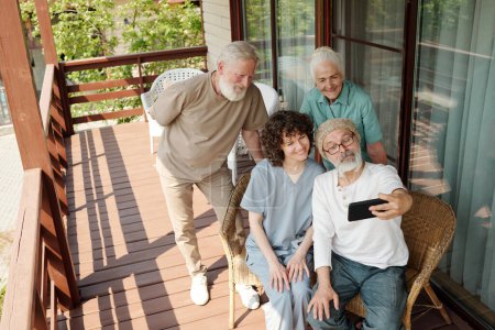 Téléchargez les photos : Les personnes âgées heureuses et leur soignant prennent selfie sur le patio de la maison de retraite tandis que l'un d'eux tenant smartphone devant leurs visages - en image libre de droit