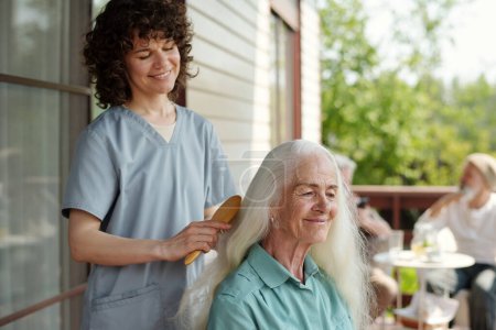 Téléchargez les photos : Heureuse jeune femme brune soignante brossant de longs cheveux blancs de patiente âgée assise devant elle sur la terrasse de la maison de retraite - en image libre de droit