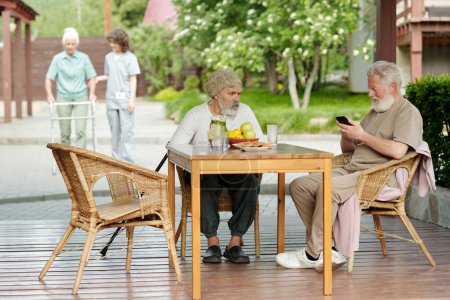 Photo for Two senior men sitting in comfortable wicker armchairs by wooden table on patio of retirement house while one of them scrolling in smartphone - Royalty Free Image