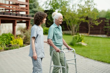 Young caregiver supporting hand of senior patient of retirement home using walker during stroll in the garden with green lawns and trees