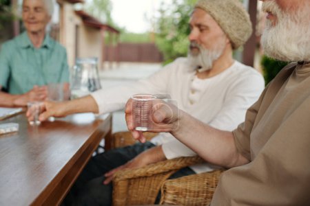 Téléchargez les photos : Focus sur la main de l'homme âgé tenant tasse transparente avec des pilules tout en étant assis à côté de la table en bois devant la caméra contre d'autres personnes âgées - en image libre de droit