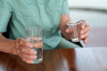 Téléchargez les photos : Mains de femme âgée en chemise bleue tenant un verre d'eau et des médicaments dans une tasse en plastique transparent assis à côté d'une table en bois - en image libre de droit