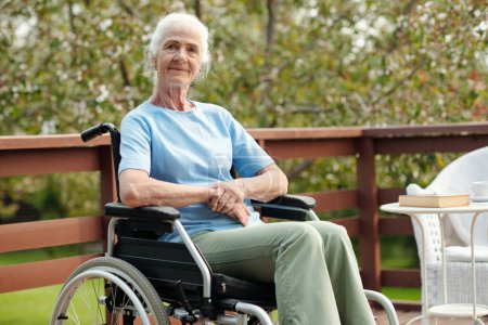 Téléchargez les photos : Femme âgée aux cheveux blancs assise en fauteuil roulant devant la caméra contre le jardin et le territoire de la maison de retraite et vous regardant - en image libre de droit