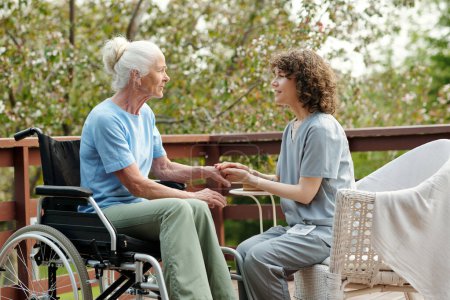 Side view of young caregiver holding hands of senior female with disability sitting in wheelchair and listening to her on terrace of retirement home