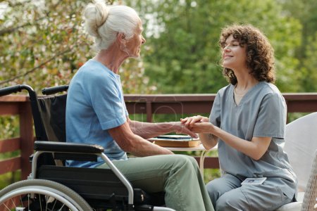 Young smiling caregiver in uniform looking at elderly woman with disability sitting in wheelchair and looking at nurse during rehabilitation treatment