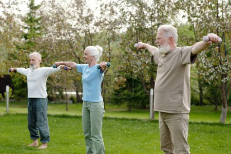 Foto de Grupo de hombres y mujeres ancianos con brazos desgarrados durante el ejercicio con pesas mientras están de pie sobre hierba verde en el jardín de la casa de retiro - Imagen libre de derechos