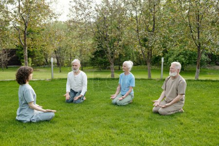 Téléchargez les photos : Trois patients âgés de la maison de retraite répéter l'exercice après leur instructeur ou soignant tout en pratiquant le yoga ensemble sur la pelouse verte - en image libre de droit