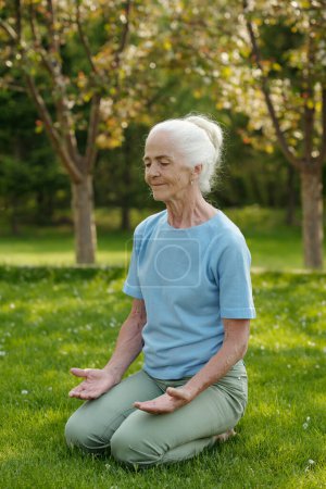 Photo for Smiling aged woman with white hair sitting in pose of lotus on green grass and practicing yoga exercises during course of rehabilitation therapy - Royalty Free Image