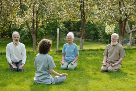 Téléchargez les photos : Jeune professeur de yoga assis sur la pelouse verte devant le groupe de trois personnes âgées vivant dans la maison de retraite pendant la formation du matin - en image libre de droit